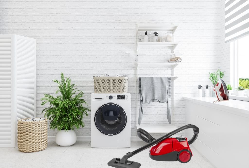 Espaço de lavanderia todo branco com  máquina de lavar roupa, aspirador de pó, ferro de passar, cestos com roupas, varal para secar a roupa e uma bela planta